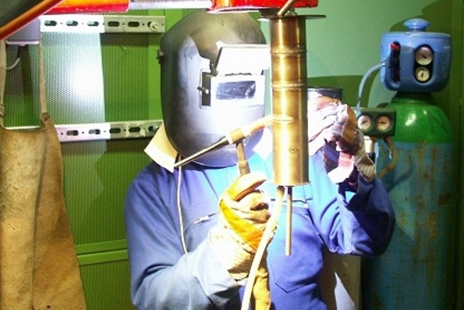 Personne équipée d'un masque de soudage, de gants de protection et d'une tenue de travail réalisant une opération de soudage sur une pièce métallique dans un atelier industriel.