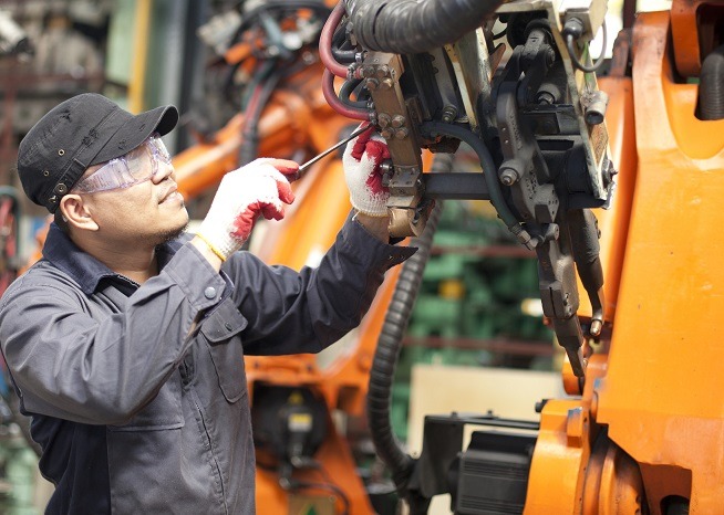 Homme portant des lunettes de protection, une casquette et des gants de sécurité intervenant avec un outil sur un équipement industriel au sein d'un atelier.