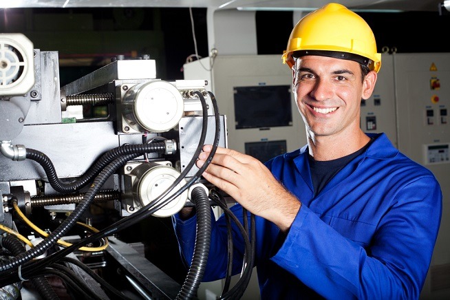 Homme portant un casque de sécurité jaune et une tenue de travail bleue intervenant sur un équipement industriel comportant des câbles, composants mécaniques et commandes techniques.