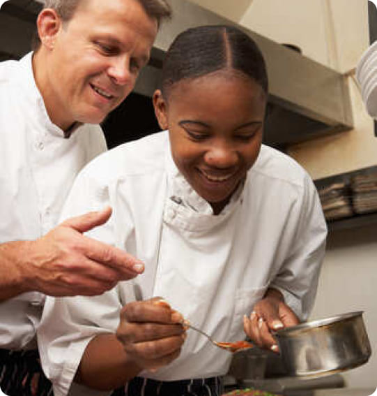 Deux personnes en tenue professionnelle de cuisine réalisent une préparation culinaire dans une cuisine. L'une utilise une cuillère tandis que l'autre l'accompagne dans le geste.