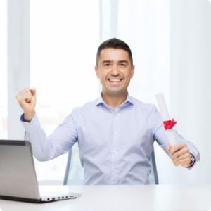 Homme assis à un bureau tenant un diplôme roulé à la main, avec un ordinateur portable posé devant lui.