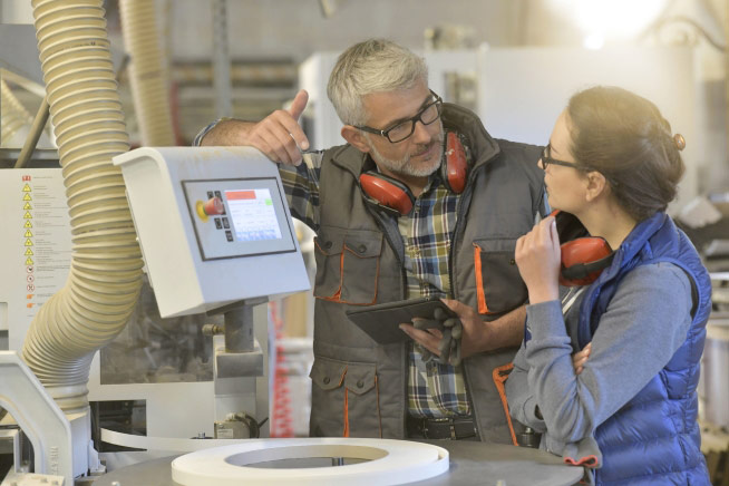 Deux personnes équipées de protections auditives échangent devant une machine industrielle. L'une tient une tablette tandis qu'un écran de commande est visible sur l'équipement.