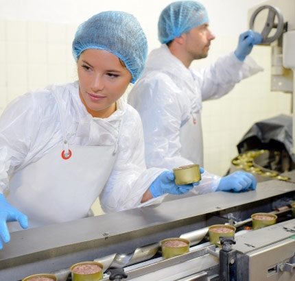 Image montrant deux personnes vêtues de tenues de protection blanches, de gants bleus et de charlottes travaillant sur une ligne de production industrielle. L'une tient une boîte métallique tandis que l'autre utilise une commande sur un équipement industriel.