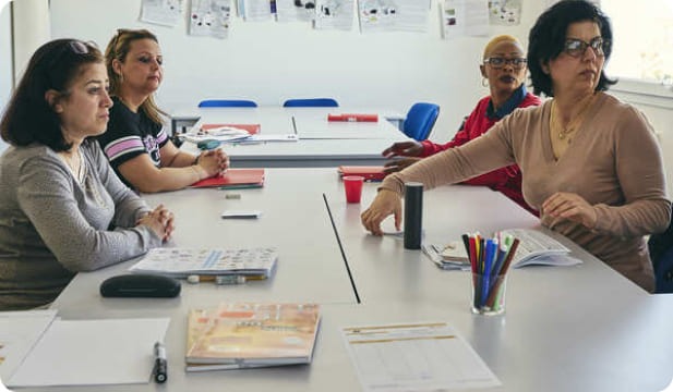 Plusieurs personnes sont assises autour d'une table lors d'une séance de formation. Des documents, stylos et supports de travail sont disposés devant elles.