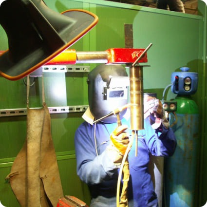 Personne équipée d’un masque de soudage et de gants de protection réalisant une opération de soudage dans un atelier industriel.