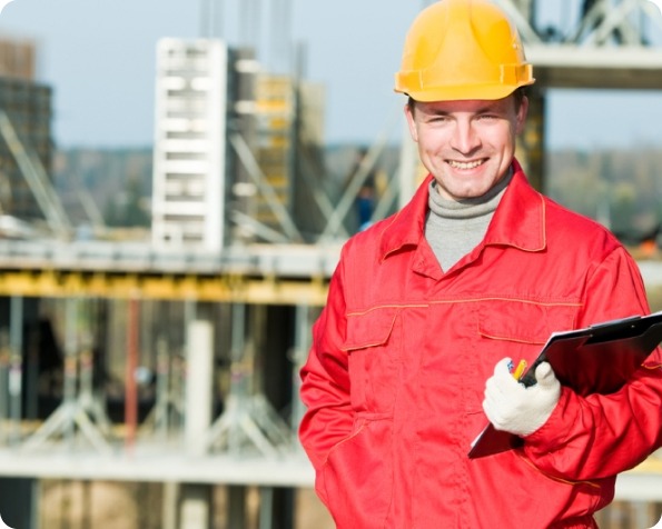 Homme portant un casque de chantier jaune et une tenue de travail rouge, tenant un classeur sur un chantier de construction.