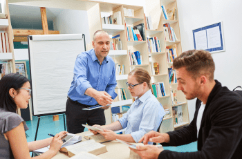 Quatre personnes réunies autour d’une table de travail. Un formateur debout en chemise bleue tend un document à une personne assise, tandis que trois autres personnes consultent des supports papier. Un tableau de présentation et une bibliothèque remplie de livres sont visibles à l’arrière-plan.