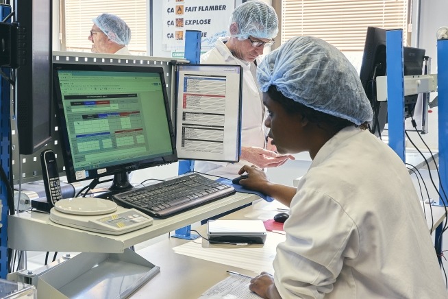 Femme en tenue de protection et charlotte utilisant un poste informatique au sein d'un environnement de production industrielle. Deux hommes équipés de protections sont visibles à l'arrière-plan.