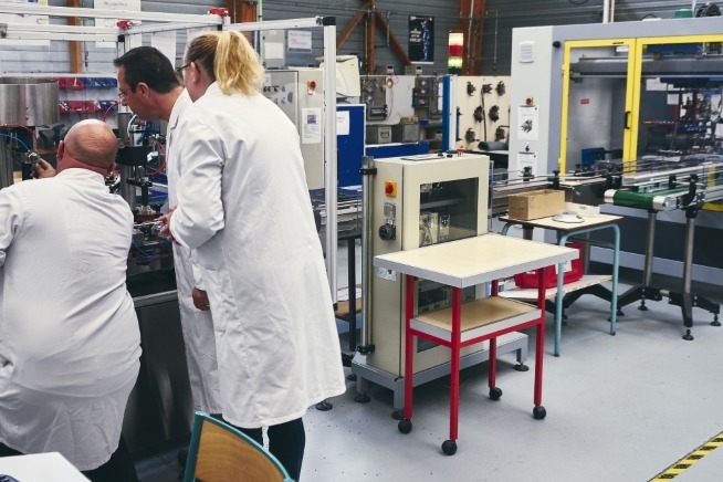 Trois personnes portant des blouses de travail blanches interviennent sur un équipement industriel dans un atelier de production. Plusieurs machines automatisées et convoyeurs sont visibles autour d'elles.