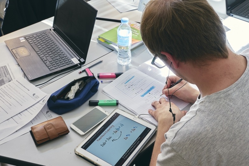 Homme assis à une table de formation prenant des notes sur un document. Un ordinateur portable, une tablette numérique, un téléphone mobile et plusieurs fournitures sont disposés sur la table.