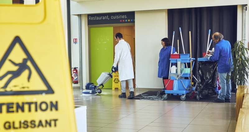 Panneau de signalisation « Attention sol glissant » au premier plan. Trois personnes réalisent des opérations de nettoyage dans un hall à l'aide d'une monobrosse et d'un chariot d'entretien.