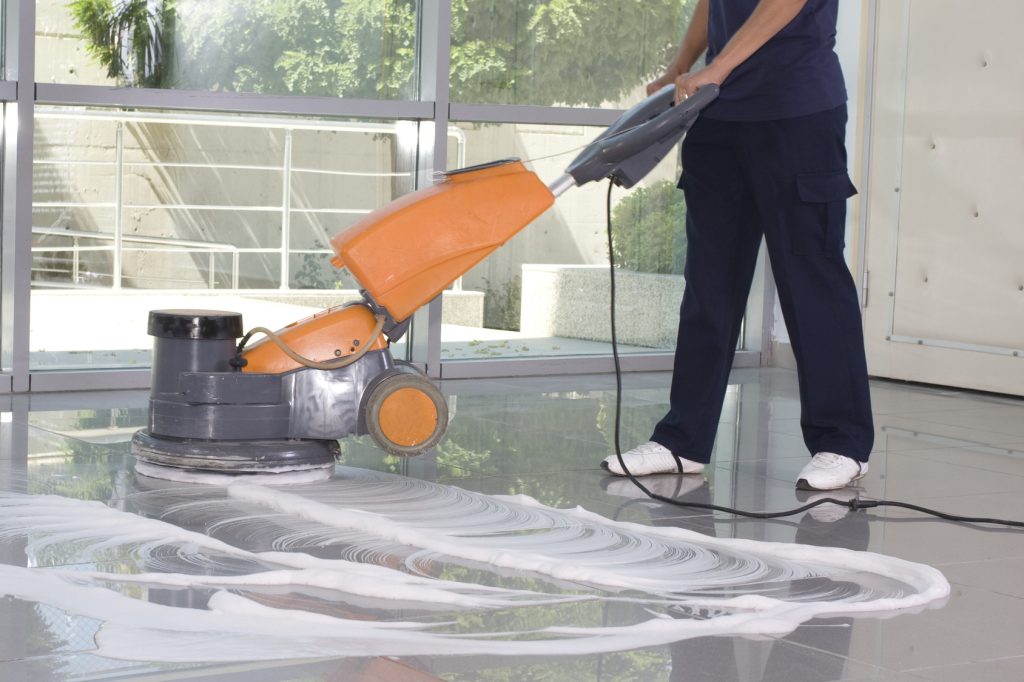 Personne utilisant une monobrosse autolaveuse pour nettoyer un sol dans un bâtiment. De la mousse est visible sur la surface en cours d'entretien.