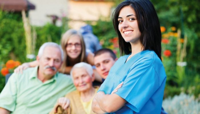 Femme en tenue médicale bleue au premier plan, avec plusieurs personnes âgées assises dans un jardin en arrière-plan.