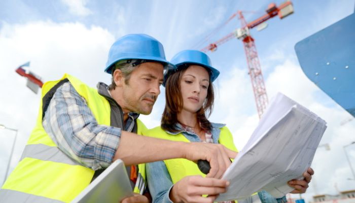 Deux personnes portant des casques de chantier bleus et des gilets haute visibilité consultent des plans sur un chantier.