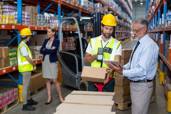 Quatre personnes dans un entrepôt logistique. Une personne portant un casque jaune et un gilet haute visibilité manipule un carton au premier plan, tandis que deux autres personnes échangent à proximité d'un chariot élévateur entre de hauts rayonnages de stockage.