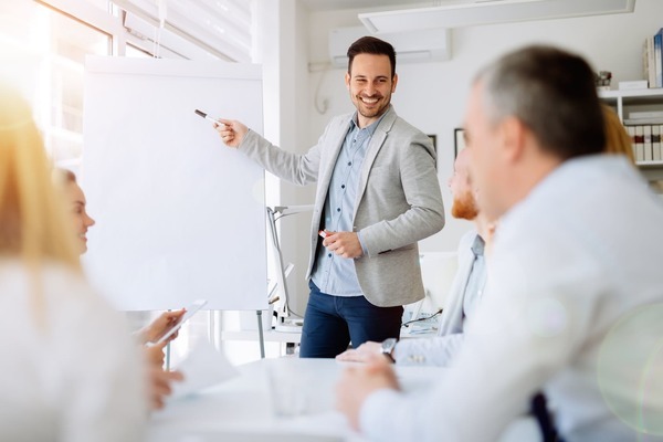 Homme debout devant un tableau de présentation blanc, pointant un élément du tableau face à plusieurs personnes assises autour d’une table de réunion dans un bureau lumineux.
