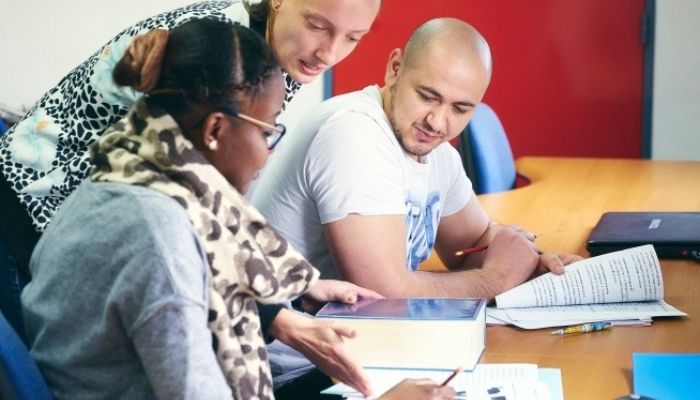 Trois personnes travaillent ensemble autour d’une table avec des documents, un livre ouvert et une tablette numérique.