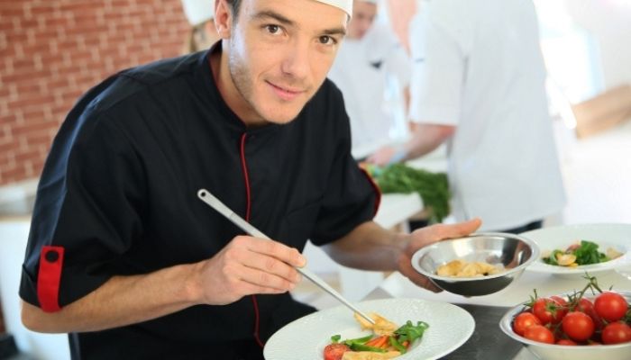Homme en tenue de cuisine tenant un bol et dressant une assiette avec une pince de précision.