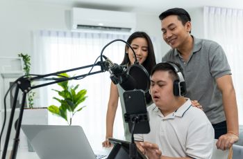 Homme portant un casque audio et parlant devant un microphone de studio, entourée de deux accompagnateurs.