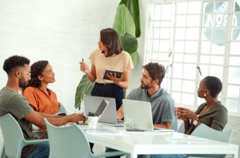Groupe de cinq personnes réunies autour d’une table de réunion avec des ordinateurs portables, tandis qu’une femme présente des informations devant le groupe.
