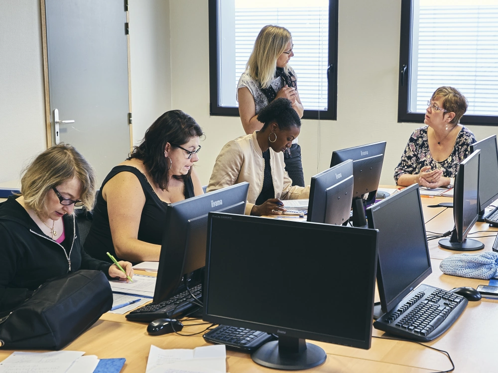 Salle de formation informatique avec plusieurs femmes assises devant des ordinateurs et une formatrice debout.