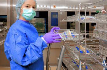 Femme portant une tenue de protection bleue, des gants violets et un masque, manipulant un plateau d’instruments dans un environnement de stérilisation.