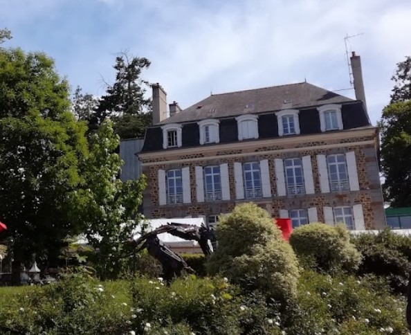 Bâtiment en pierre de plusieurs étages avec volets blancs, entouré d’arbres et d’espaces verts.