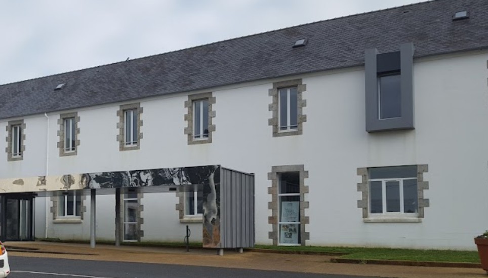 Bâtiment blanc à deux étages avec encadrements de fenêtres en pierre et auvent métallique à l’entrée.