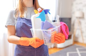 Personne portant des gants de protection orange et un tablier, tenant un bac contenant des produits et accessoires de nettoyage.