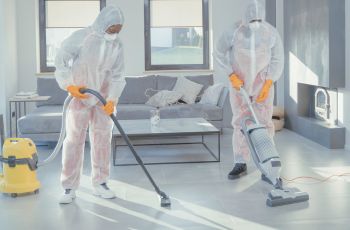Deux personnes en combinaison de protection blanche utilisent des aspirateurs professionnels dans une pièce.