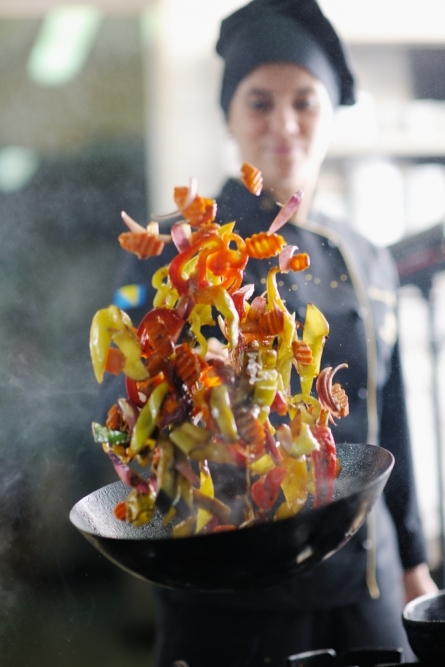 Poêlée de légumes colorés projetés en l'air au-dessus d'un wok. Une femme en tenue de cuisine est visible à l'arrière-plan.