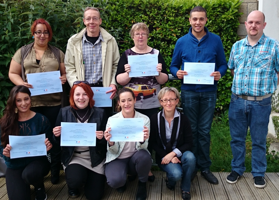 Groupe de neuf personnes posant en extérieur devant une haie végétale. Plusieurs personnes tiennent un certificat ou un diplôme devant elles.