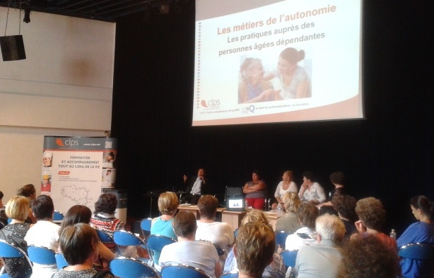 Personnes assises dans une salle de conférence face à une scène. Une présentation est projetée sur un grand écran intitulée « Les métiers de l'autonomie - Les pratiques auprès des personnes âgées dépendantes ».