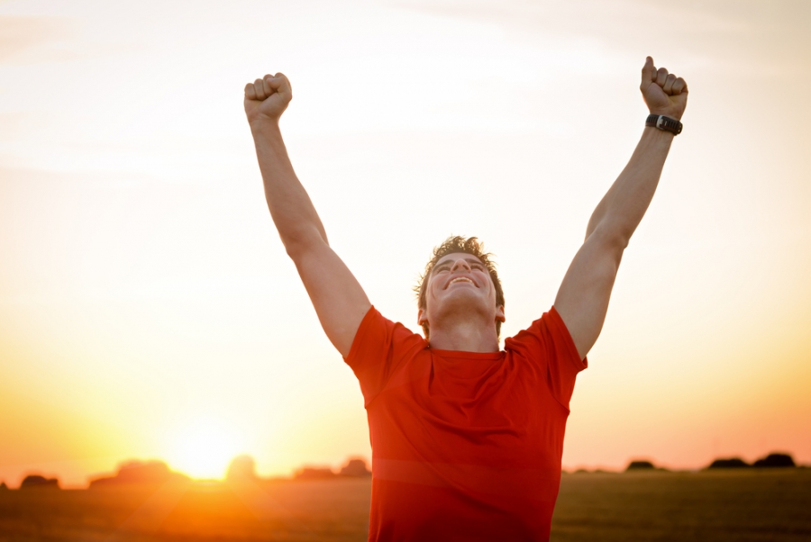 Homme portant un tee-shirt rouge levant les deux bras vers le ciel dans un paysage au coucher du soleil.