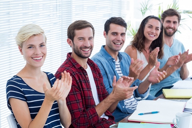 Groupe de cinq personnes assises autour d'une table en salle de réunion, applaudissant et souriant face à l'objectif.
