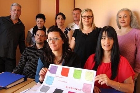 Groupe de huit personnes réunies dans une salle autour d'une table. Deux personnes au premier plan présentent un document coloré composé de plusieurs encadrés.