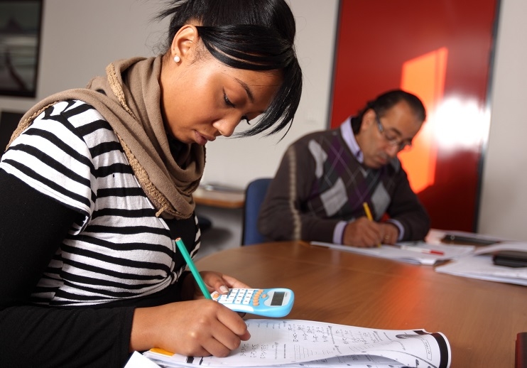 Deux femmes sont assises à une table et remplissent des documents. L'une utilise une calculatrice tandis que des feuilles d'exercices sont disposées devant elle.