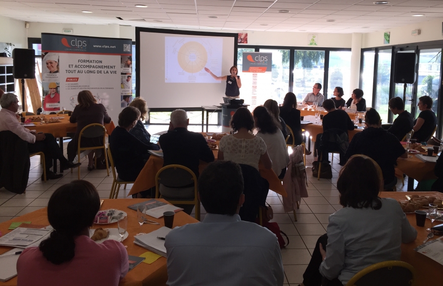 Participants assis autour de tables lors d'une réunion ou d'une présentation. Une intervenante présente un diaporama projeté sur un écran.