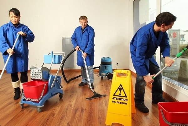 Trois personnes en blouse de travail bleue réalisent des opérations de nettoyage dans une pièce. Un aspirateur, un chariot de ménage, des seaux et un panneau « Attention sol glissant » sont visibles.
