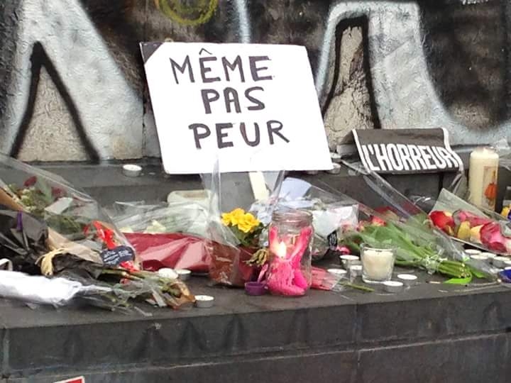 Panneau portant l’inscription « MÊME PAS PEUR » entouré de bouquets de fleurs, de bougies et de messages déposés lors d’un hommage.