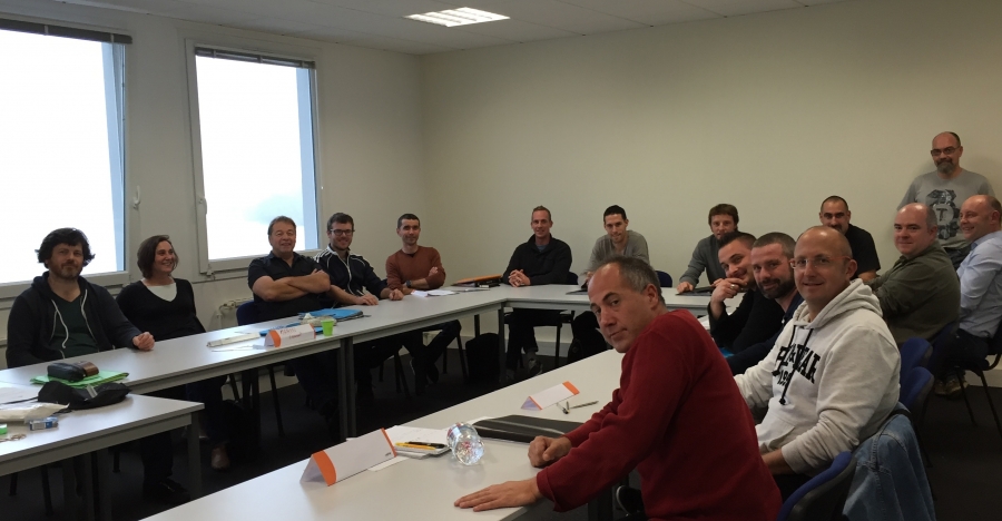 Groupe de personnes assises autour de tables disposées en U dans une salle de formation. Un homme est debout au fond de la salle.