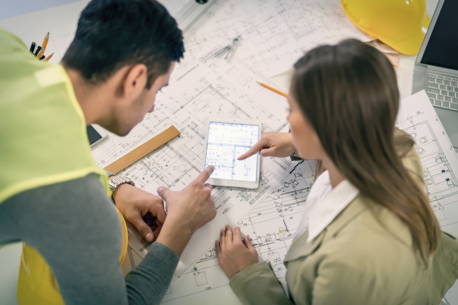 Un homme et une femme consultent des plans de construction et une tablette numérique posés sur une table de travail. Un casque de chantier jaune est visible à proximité.