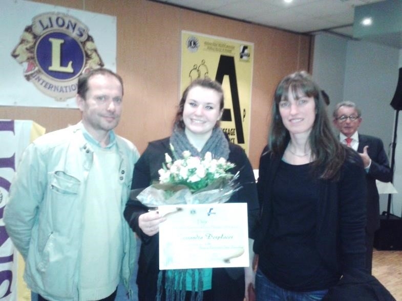 Trois personnes posent côte à côte lors d'une cérémonie de remise de prix. La personne au centre tient un bouquet de fleurs et un diplôme.