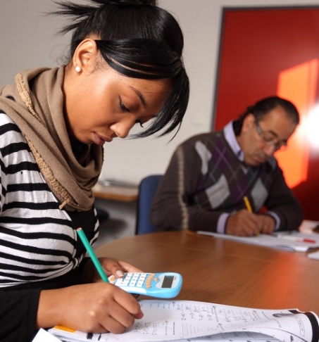 Femme écrivant sur une feuille d’exercices avec un crayon tout en utilisant une calculatrice de poche, assise à une table de formation. Un homme travaille également à l’arrière-plan.