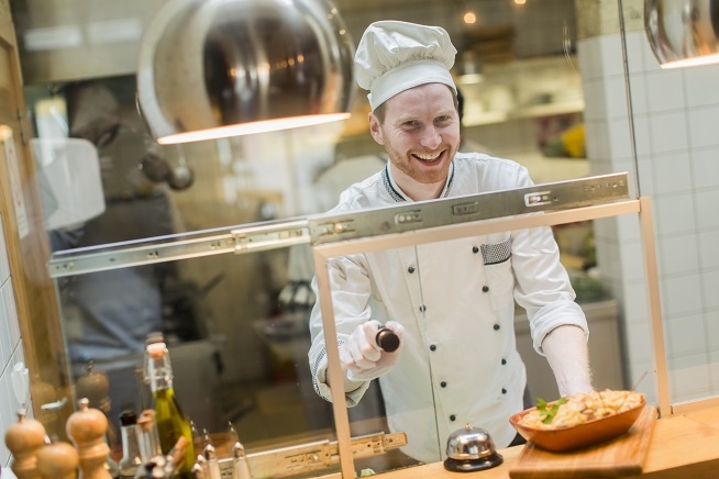 Homme en tenue de cuisine blanche et toque de chef travaillant derrière un passe-plat dans une cuisine professionnelle.