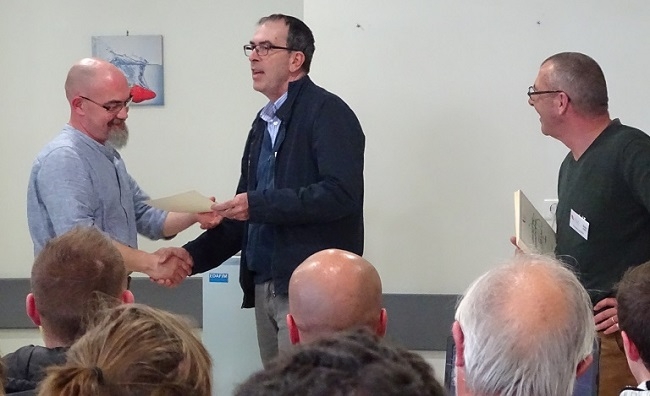 Trois hommes participent à une remise de diplôme dans une salle de réunion. L'un remet un document à un autre tandis qu'un troisième tient un certificat.
