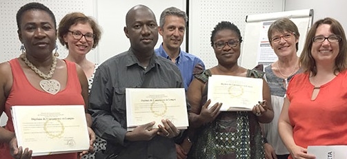 Groupe de sept personnes posant côte à côte dans une salle, plusieurs tenant un diplôme devant elles.