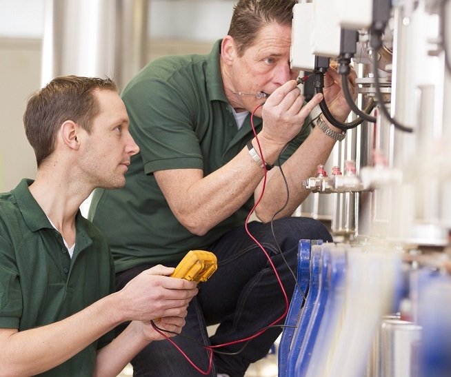 Deux hommes en tenue de travail verte interviennent sur une installation industrielle. L'un utilise un appareil de mesure tandis que l'autre observe l'équipement.