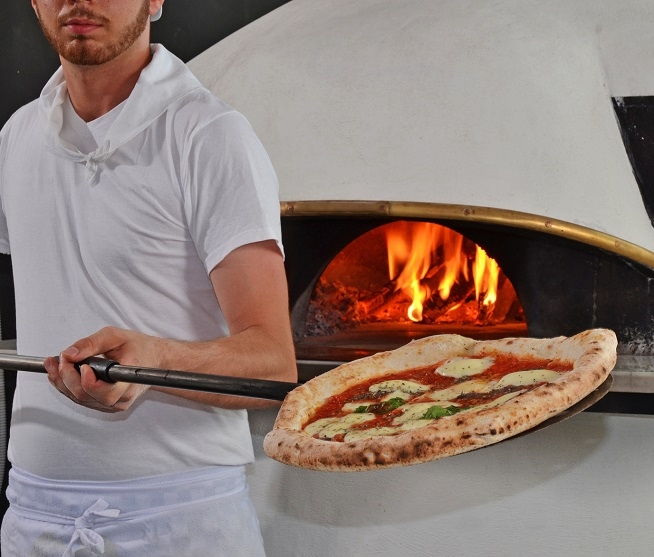 Homme en tenue de cuisine tenant une pelle à pizza devant un four à bois. Une pizza garnie est visible au premier plan.