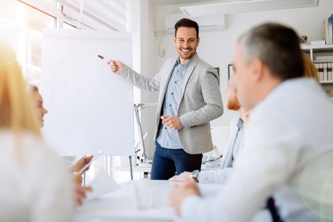 Homme debout devant un paperboard lors d’une réunion de travail, face à plusieurs participants assis autour d’une table.